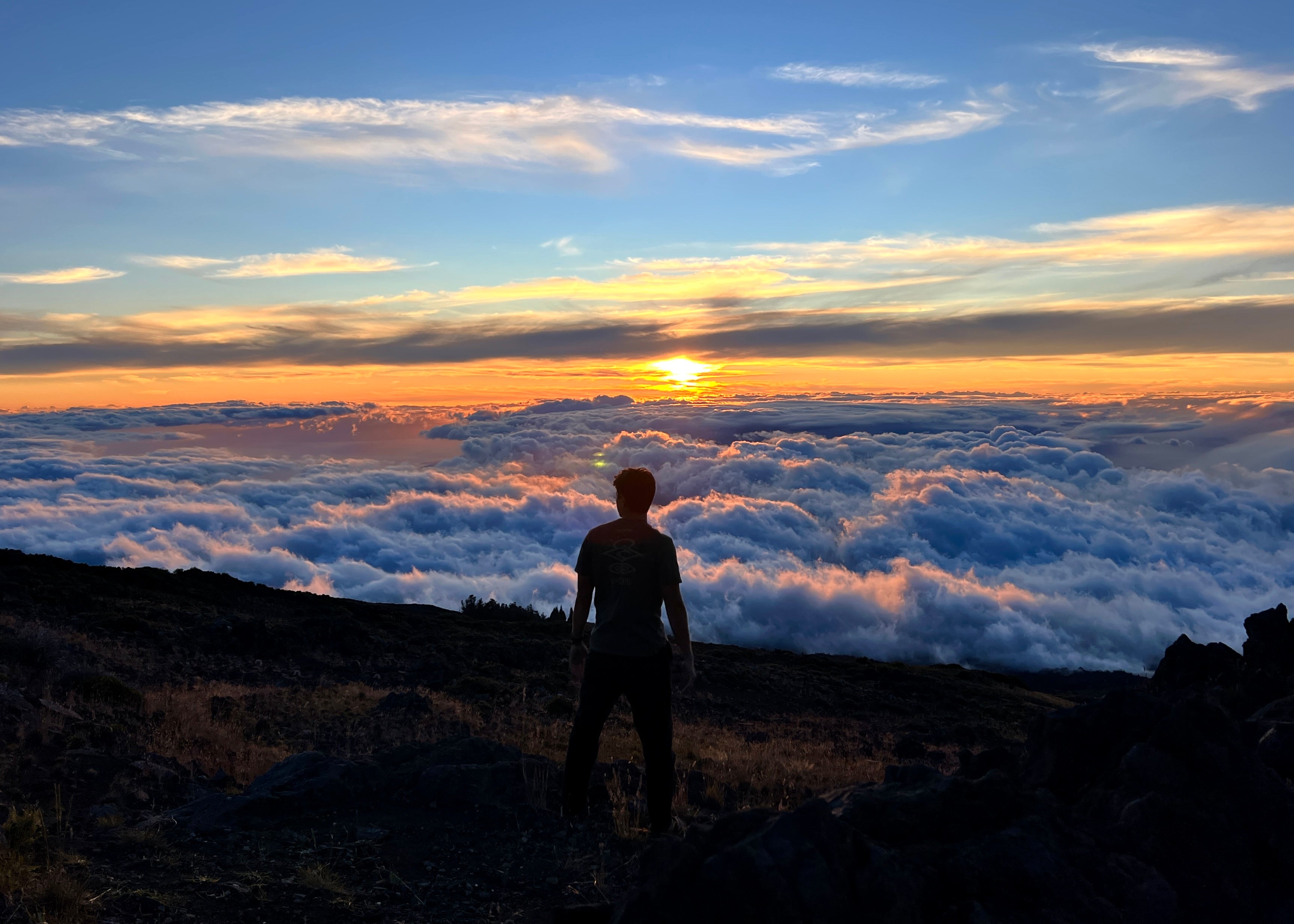 Person standing on a mountain top looking at a sunset over clouds